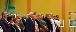Zaproszeni goście i przedstawiciele instytucji stoją podczas oficjalnej części uroczystości nadania Szkole Podstawowej w Dąbrówce Dużej imienia Parku Krajobrazowego Wzniesień Łódzkich. W tle widoczne są fotografie prezentujące walory przyrodnicze parku