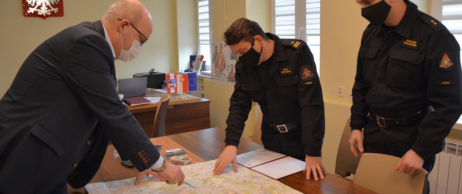Starosta turecki w raz z komendantem powiatowy wraz z strażakiem pochylają się nad mapą powiatu.