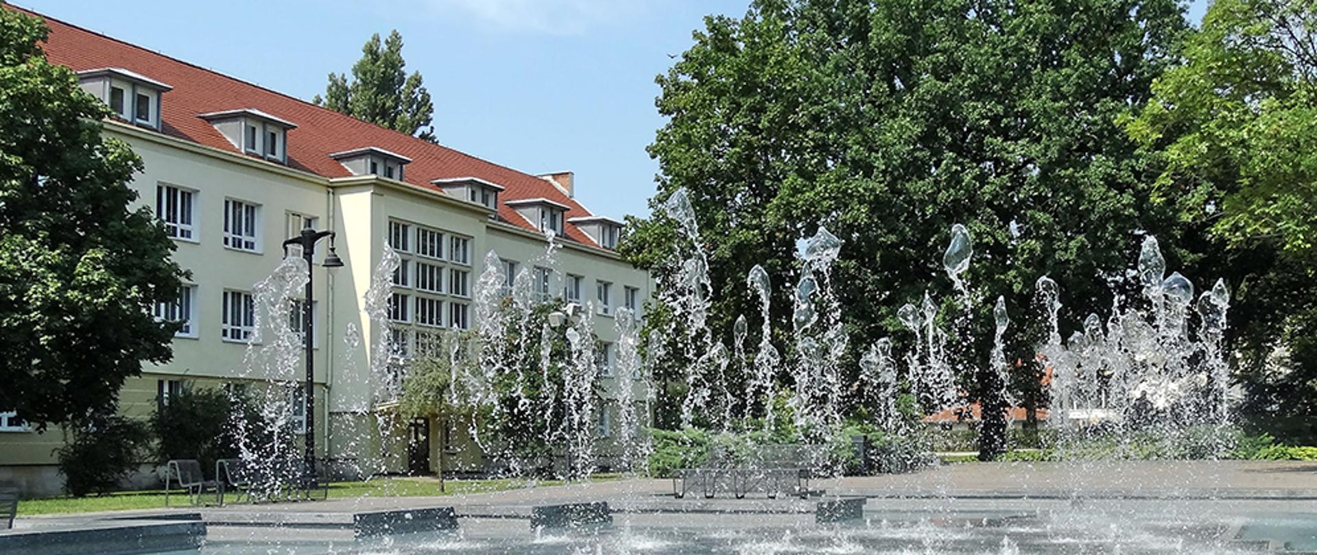 Zdjęcie przedstawia budynek szkoły w otoczeniu fontanny