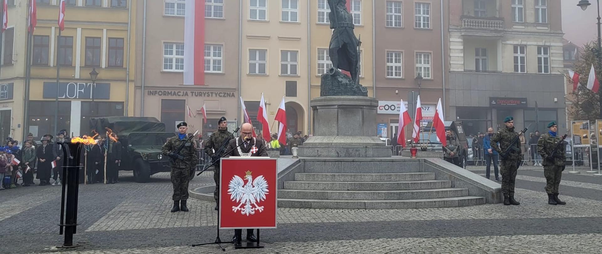 Na zdjęciu jest przedstawiony Prezydent Miasta Grudziądza stojący za mównicą i przemawiający do zgromadzonych. W tle znajduje się pomnij Żołnierza Polskiego i zgromadzeni goście. 