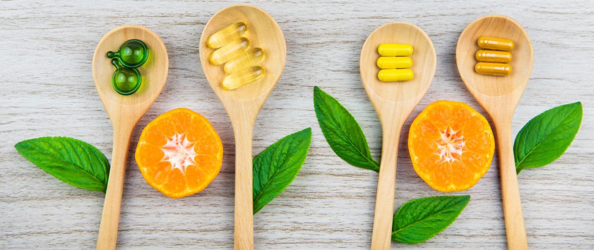 Na grafice widzimy 4 drewniane łyżki w których znajdują się tabletki, miedzy łyżkami znajdują się zielone listki oraz połówki pomarańczowego owocu. 