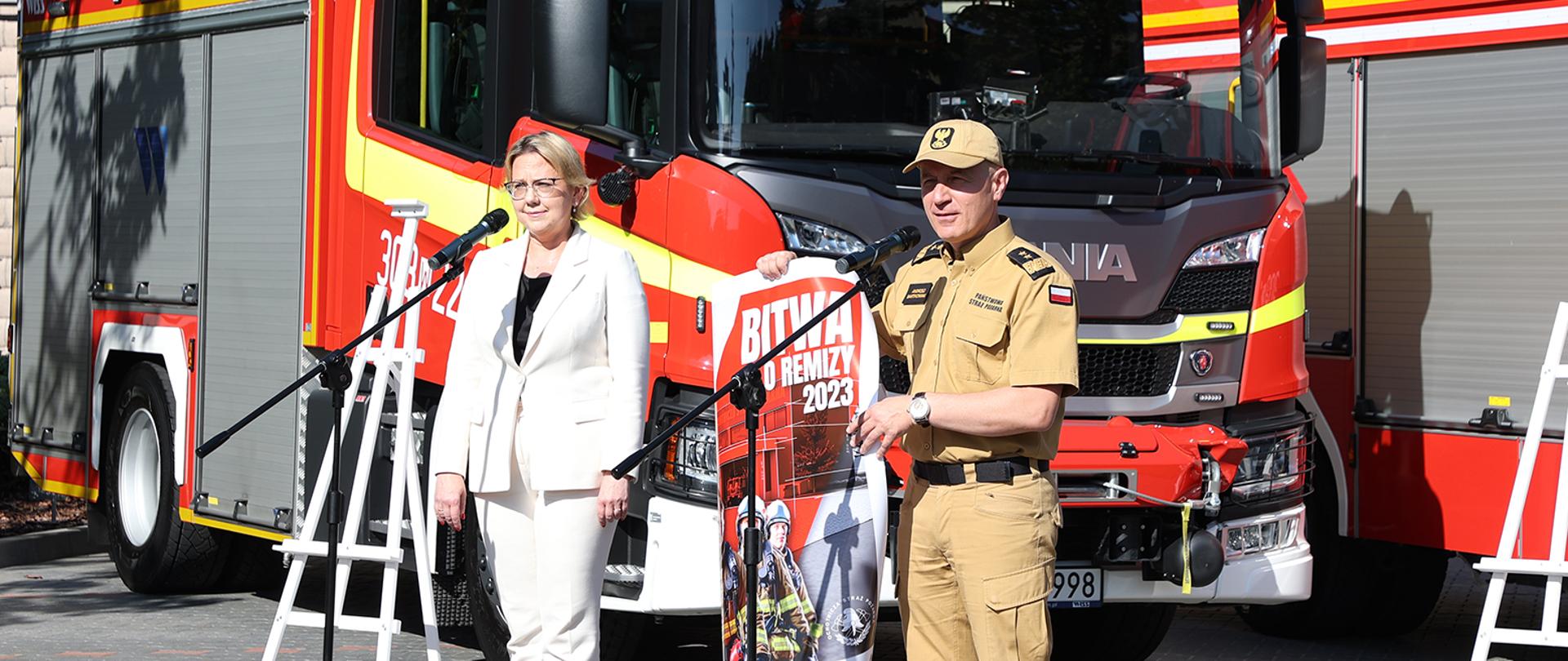 Na zdjęciu po lewej stronie minister klimatu i środowiska Pani Anna Moskwa, na zdjęciu po prawej stronie Gen. brygadier Andrzej Bartkowiak komendant główny PSP w trakcie konferencji, stoją przy mikrofonach, prezentują plakat reklamujący „Bitwę o remizy”. Za nimi w tle znajduje się czerwony samochód ratowniczo-gaśniczy.