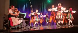 W pomieszczeniu - sala widowiskowa Ośrodka Kultury w Oławie. Na scenie, na tle czarnych kotar członkowie Zespołu Tańca Długołęka tańczą w strojach ludowych. 