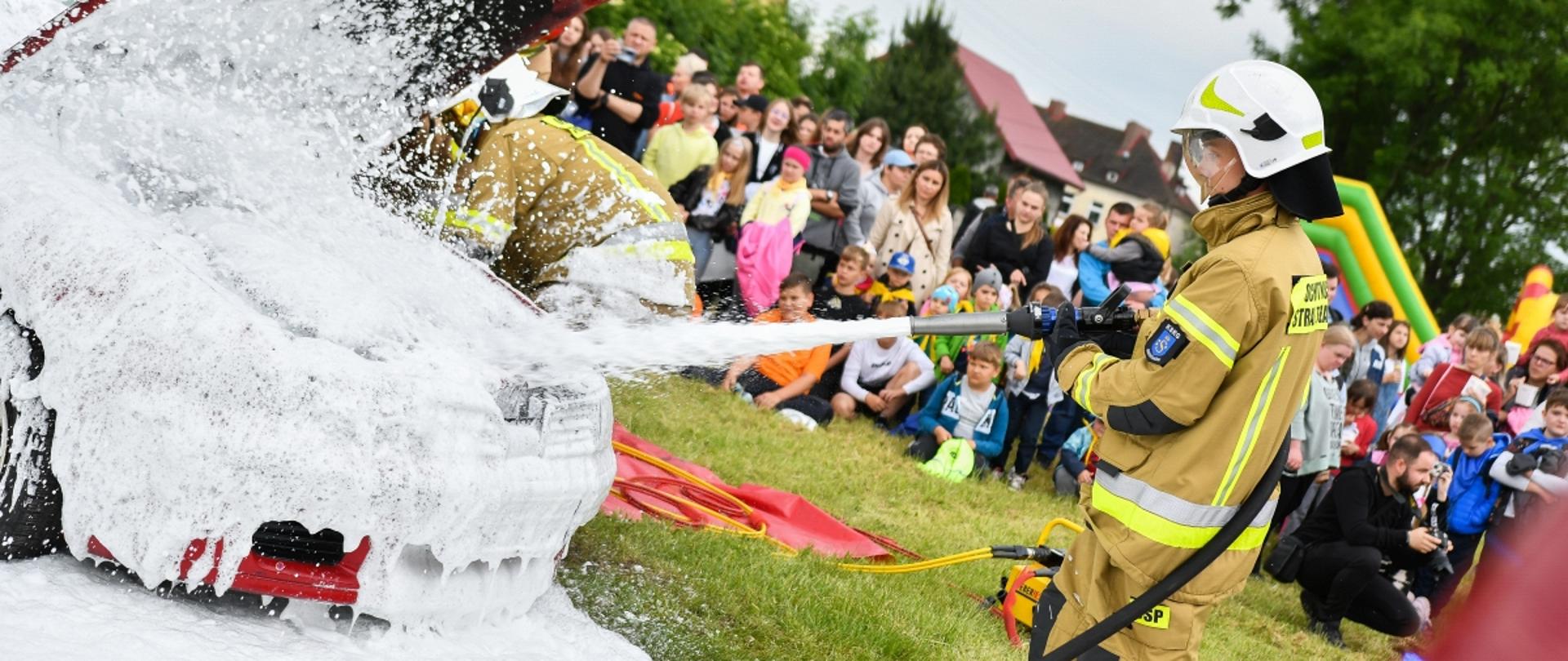 Pokazy strażackie na festynie z okazji Dnia Dziecka, na pierwszym panie strażak w ubraniu bojowym gasi pianą samochód na boisku, w tle uczniowie i osoby dorosłe oglądające pokaz