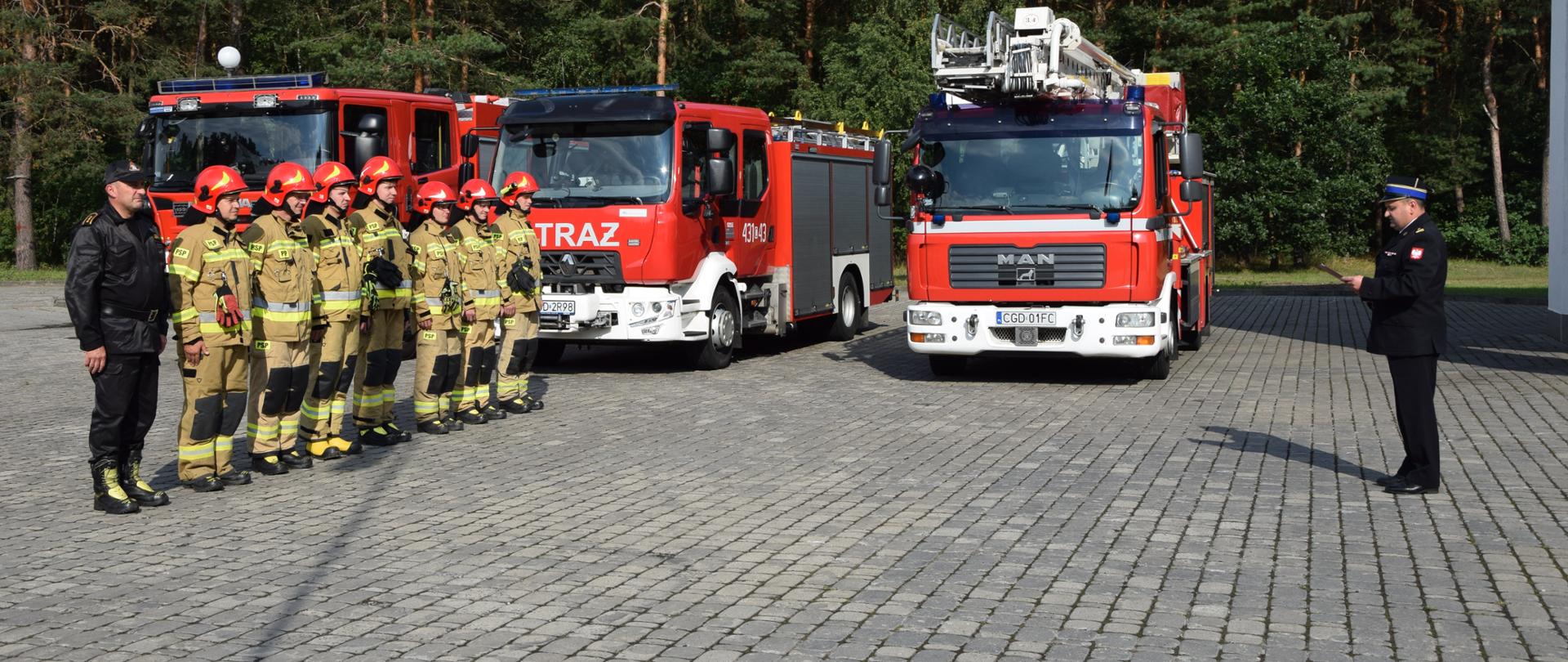 Strażacy w ubraniach specjalnych ustawieni w szeregu, stoją ustawieni na tle wozów bojowych. Obok nich w szeregu stoi strażak w umundurowaniu dowódczo-sztabowym. Przed nimi stoi Komendant Powiatowy z bordowa teczką w dłoniach i wygłasza przemówienie.