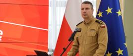 Na zdjęciu znajduje się mężczyzna w mundurze Państwowej Straży Pożarnej, stojący przy drewnianej mównicy i wygłaszający przemówienie. Na jego mundurze widnieje naszywka z imieniem i nazwiskiem oraz oznaczenie PSP. W tle znajdują się flagi Polski i Unii Europejskiej, a także ekran z czerwonym tłem i białymi elementami graficznymi.