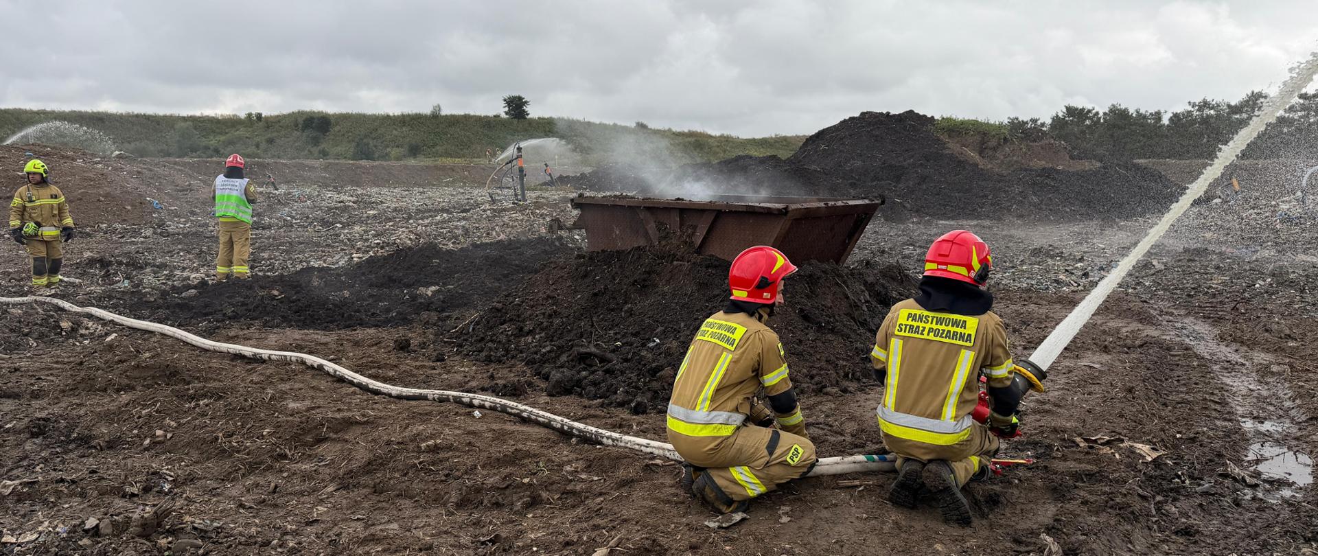 Strażacy używają wężów strażackich na terenie wysypiska pokrytego ciemną ziemią. Powyżej pochmurne niebo. 