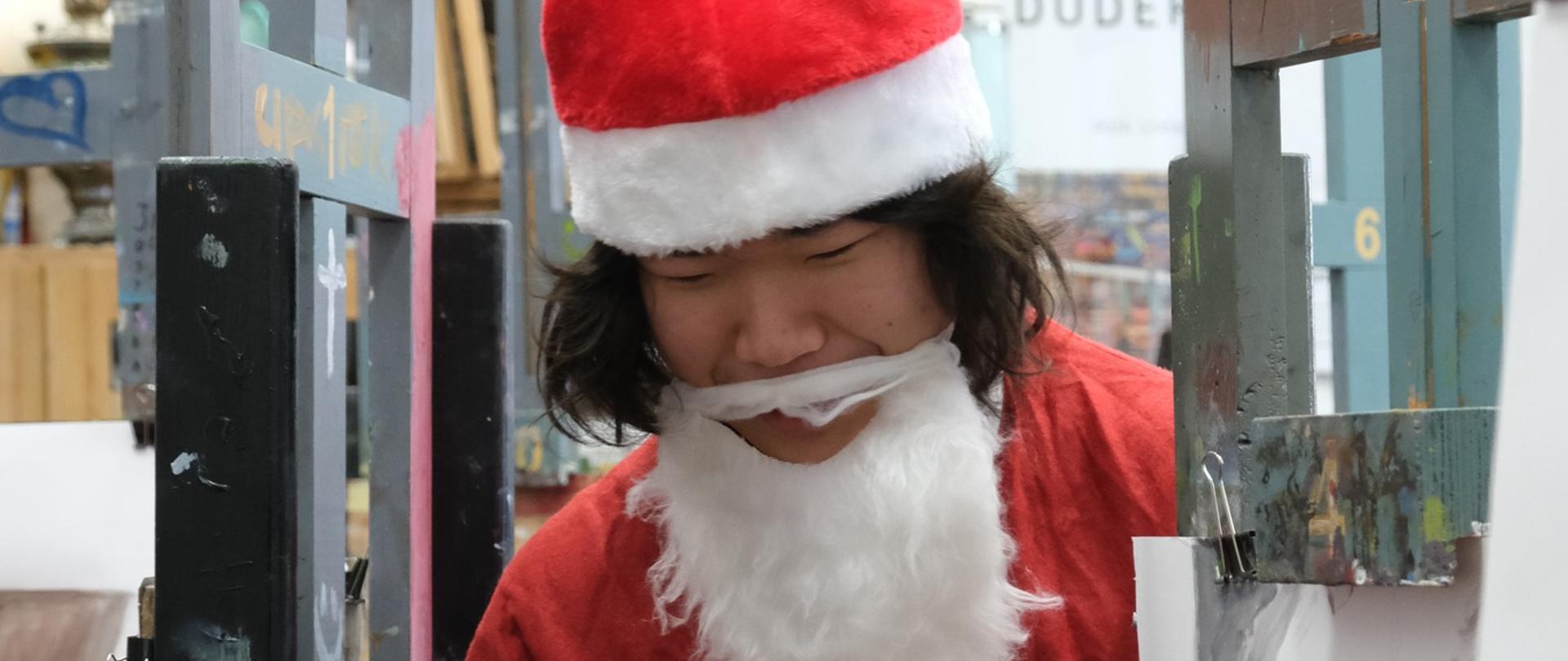 Młody człowiek przebrany za Świętego Mikołaja trzyma worek ze słodyczami. Na pierwszym planie siedząca tyłem dziewczyna zagląda do worka.