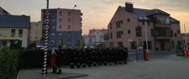 Zdjęcie przedstawia strażaków podczas uroczystej zmiany służby.
W tle budynki.

