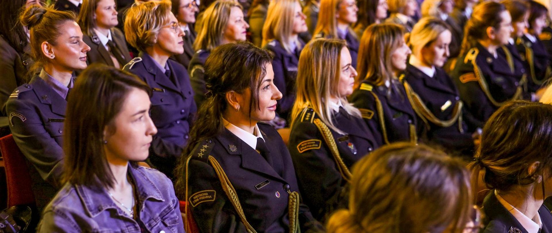 Na zdjęciu widzimy kobiety w mundurach różnych formacji siedzące na widowni. Na pierwszym planie kobieta w stopniu kapitana Państwowej Straży Pożarnej.