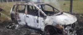 Na drodze gruntowej prowadzącej w kierunku lasu został podpalony samochód osobowy marki Mazda. Na zdjęciu widać całkowicie zniszczony w wyniku pożaru wrak samochodu stojący przy ogrodzeniu z siatki leśnej. 