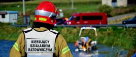 Na pierwszym planie widoczny jest strażak stojący tyłem do obiektywu, ubrany w specjalną kamizelkę z napisem „KIERUJĄCY DZIAŁANIEM RATOWNICZYM”. Obserwuje on akwen wodny, na którym operuje łódź ratownicza z załogą. W tle przy drodze zaparkowane są czerwone pojazdy ratownicze.