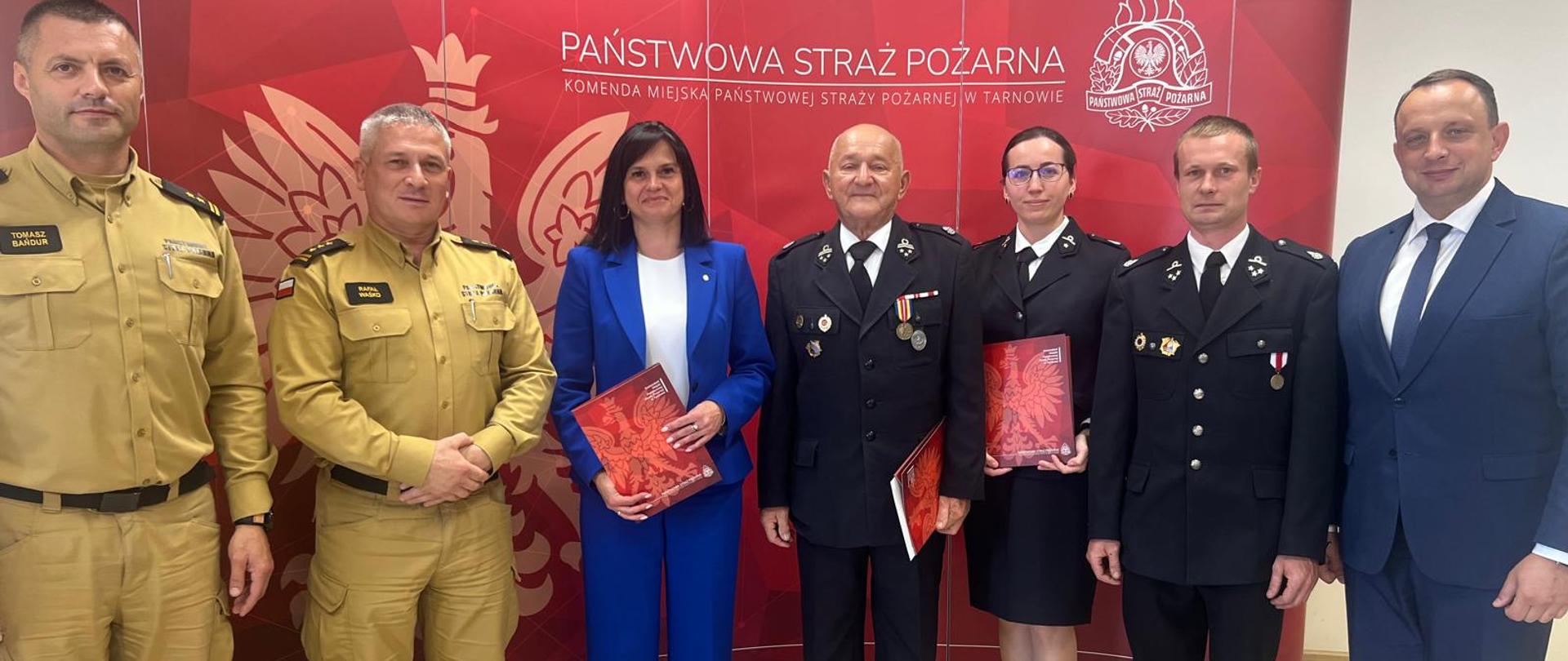 na tle czerwonej ścianki Państwowej Straży Pożarnej stoją strażacy oraz przedstawiciele samorządu. Strażacy PSP ubrani w piaskowe mundury, strażacy OSP w granatowe mundury