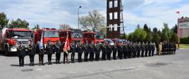 Na zdjęciu widać pododdział strażaków stojących na placu przed komendą. Wszyscy ubrani są w umundurowanie wyjściowe. W tle widoczne są ustawione samochody pożarnicze.