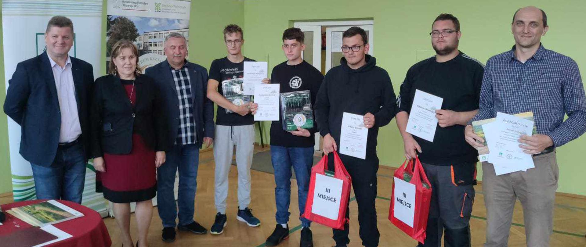 Grupa mężczyzn i jedna kobieta stoją w sali gimnastycznej podczas ceremonii wręczenia nagród. Czterej młodzi mężczyźni trzymają dyplomy i czerwone torby z napisem „III MIEJSCE”. W tle widoczne są banery Sieci Szkół Rolniczych.
