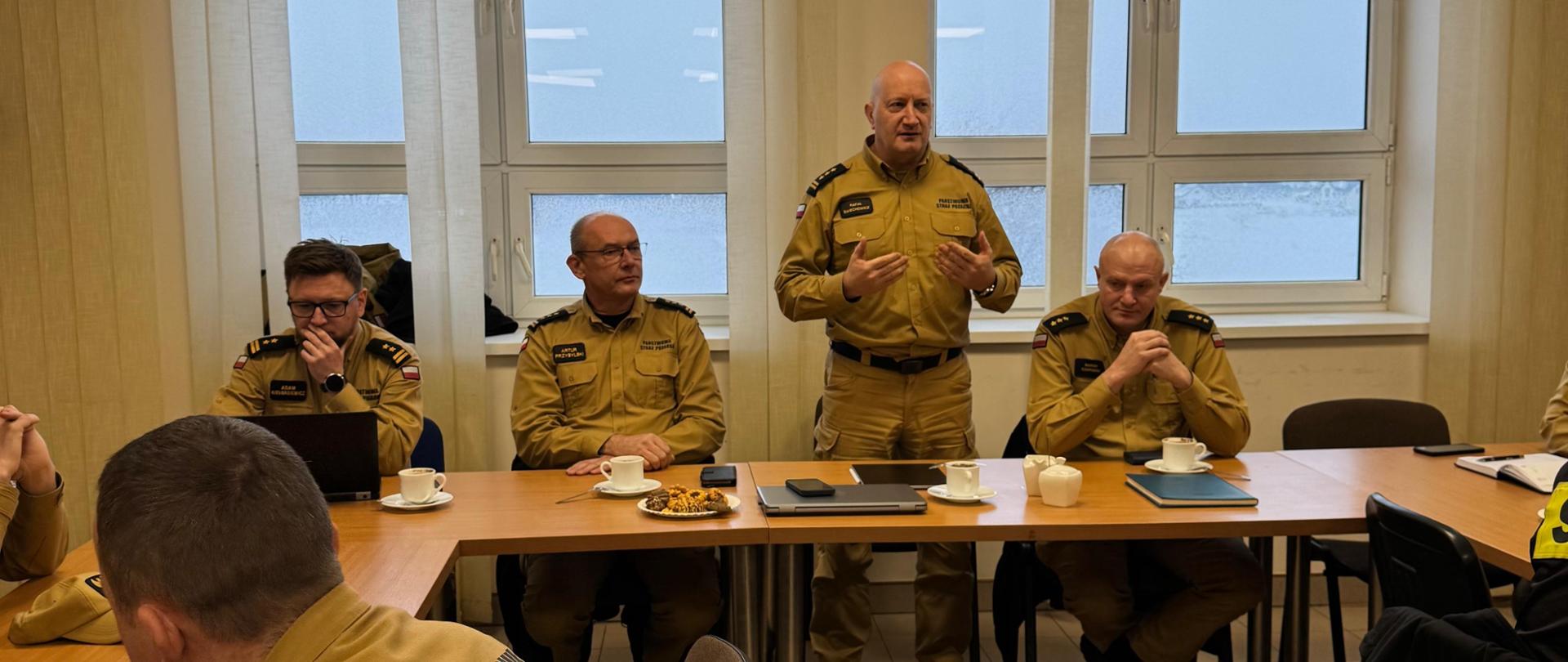 Na pierwszym planie widać przedstawicieli kierownictwa Państwowej Straży Pożarnej: od lewej bryg. Adam Kiełbasiewicz – Naczelnik Wydziału Organizacji i Nadzoru Komendy Wojewódzkiej PSP w Toruniu, st. bryg. Artur Przybylski – Zastępca Kujawsko-Pomorskiego Komendanta Wojewódzkiego PSP, st. bryg. Rafał Świechowicz – Kujawsko-Pomorski Komendant Wojewódzki PSP oraz kpt. Marcin Czarnecki – Zastępca Komendanta Powiatowego PSP w Rypinie. Funkcjonariusze ubrani są w umundurowanie służbowe. W tle widoczne są duże okna budynku.