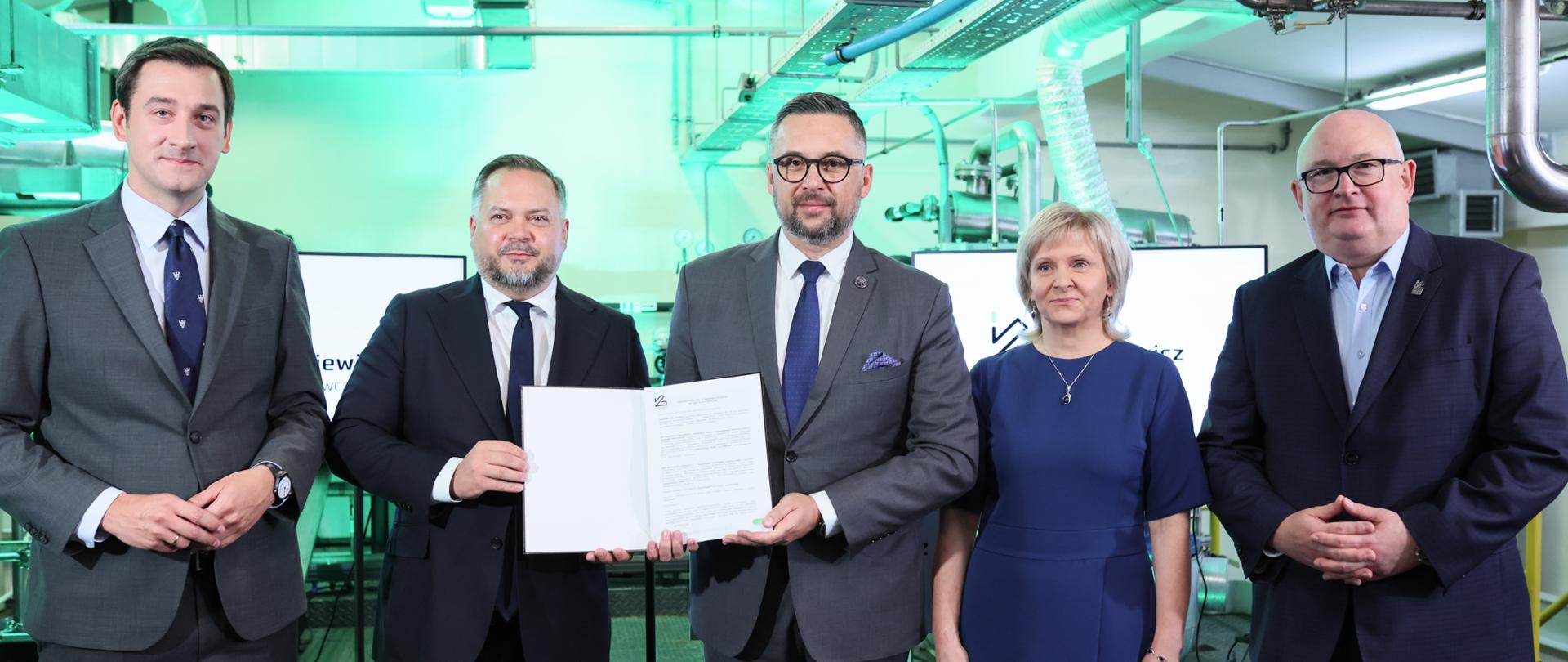 Sieć Badawcza Łukasiewicz wzmacnia suwerenność Polski w produkcji materiałów wybuchowych i amunicji - na zdjęciu 5 osób - czterech mężczyzn i kobieta. śród nich Minister Marcin Kulasek trzymający dokument z logo Łukasiewicz.