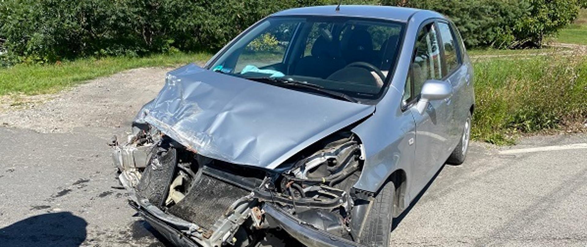 Rozbity srebrny samochód osobowy stojący na jezdni.W tle krzaki i czerwony dach budynku. Uszkodzony cały przód. Ciemna plama pod pojazdem rozbitych płynów eksploatacyjnych. 