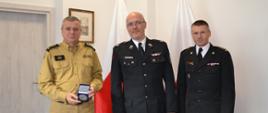 Pomorski komendant wojewódzki Państwowej Straży Pożarnej trzyma medal w obecności druha ochotniczej straży pożarnej oraz komendanta powiatowego Państwowej Straży Pożarnej w Pucku.