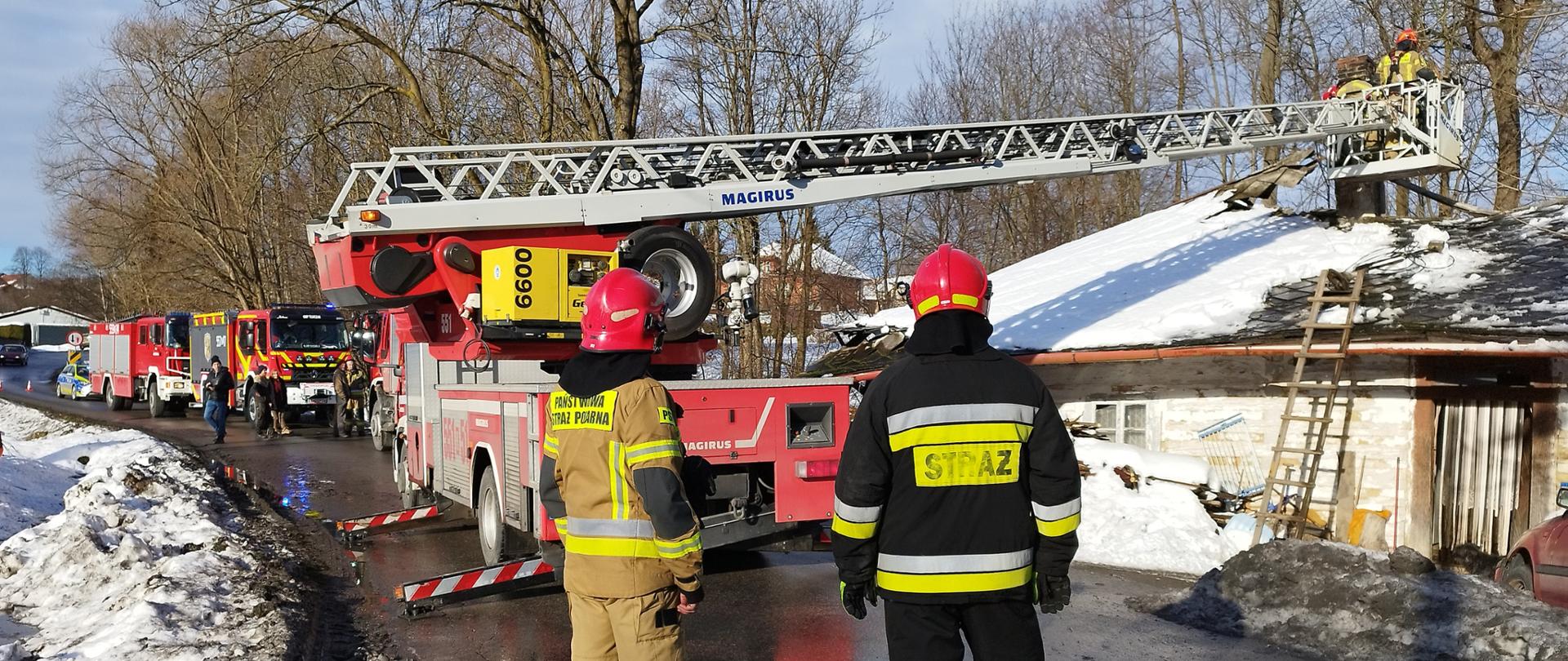 Na pierwszym planie dwóch strażaków widocznych od tyłu, w tle po prawej stronie budynek mieszkalny z zawalonym dachem, po lewej stronie autodrabina i kolejne samochody PSP i OSP.