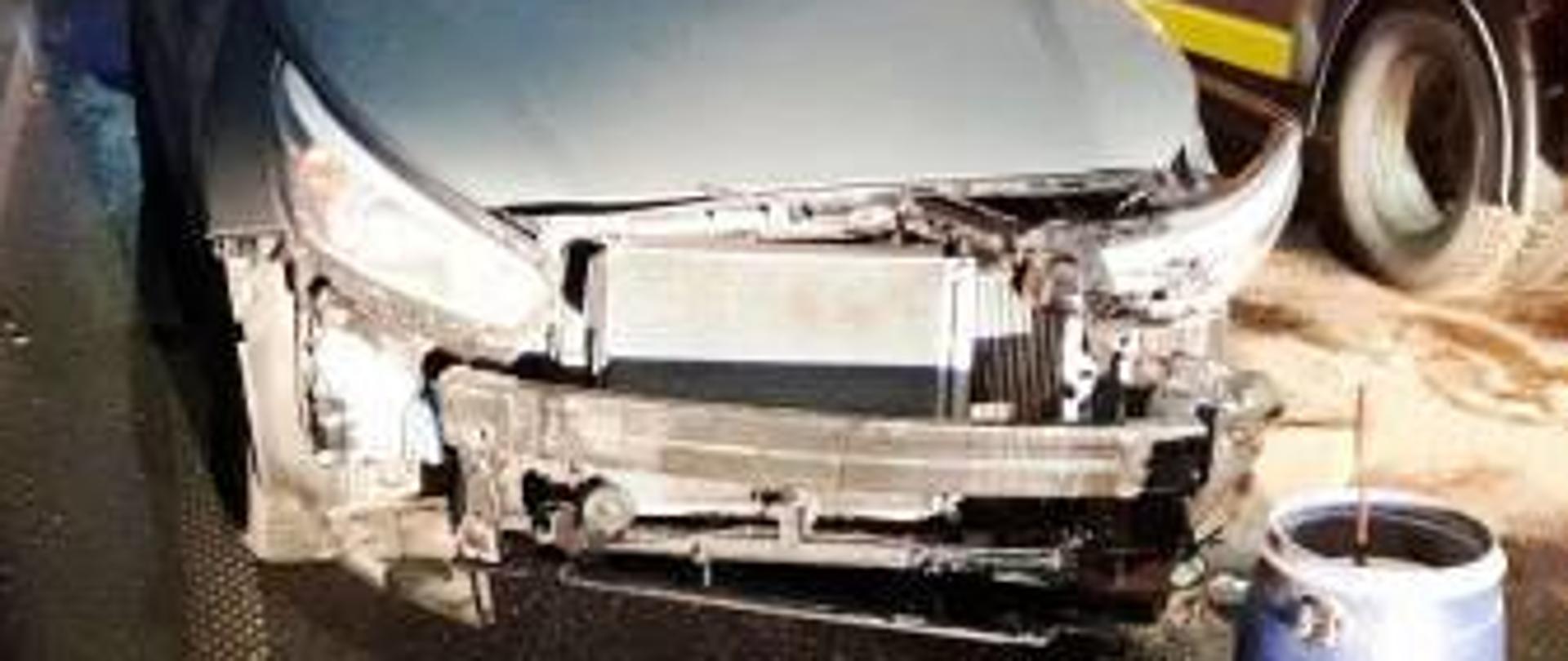 Widok przodu rozbitego samochodu. Obok stoi laweta. Z przodu rozbitego samochodu beczka z sorbentem.