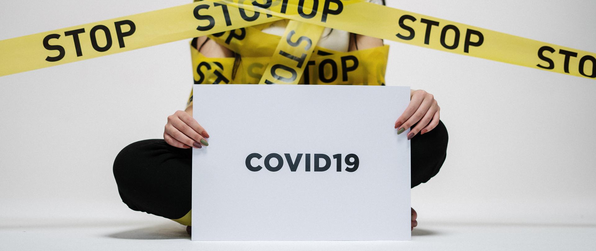 Sylwetka osoby siedzącej po turecku, w dłoniach kartka z napisem COVID19