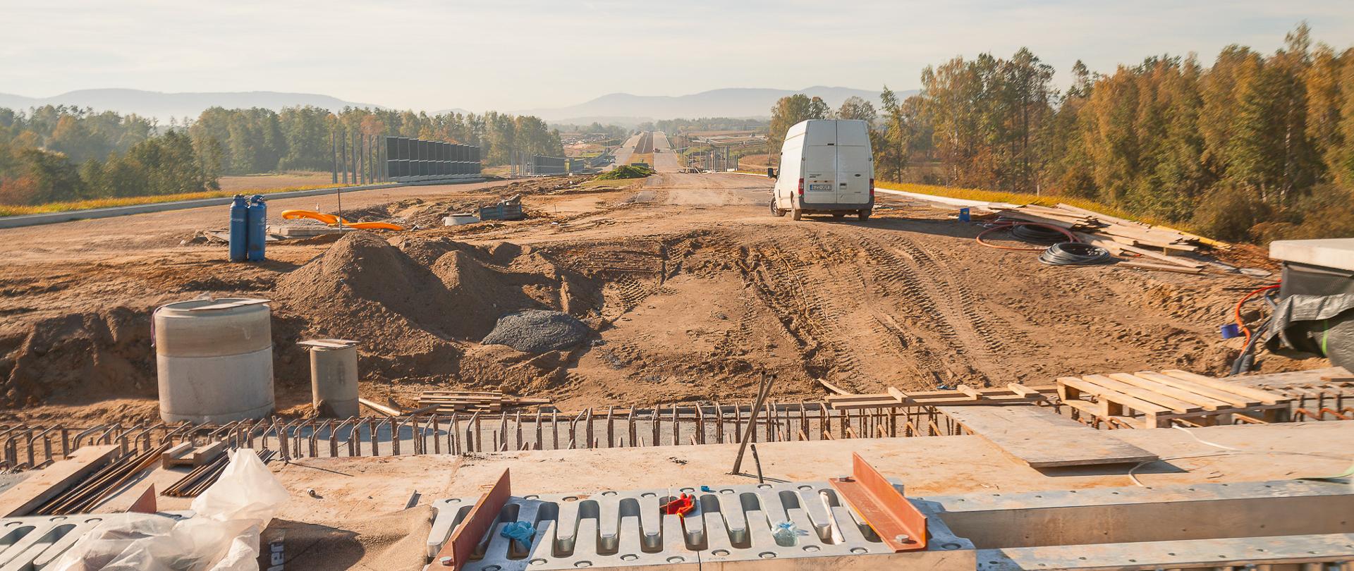 Zdjęcie przedstawia drogę ekspresową w budowie. Na pierwszym planie widoczne są elementy konstrukcyjne, takie jak metalowe belki i drewniane deski. W tle znajduje się rozległy teren budowy z kopcami ziemi, sprzętem budowlanym oraz pojazdami. Po prawej stronie widoczny jest biały samochód dostawczy. Całość otoczona jest drzewami i roślinnością, a w oddali można dostrzec góry na horyzoncie.