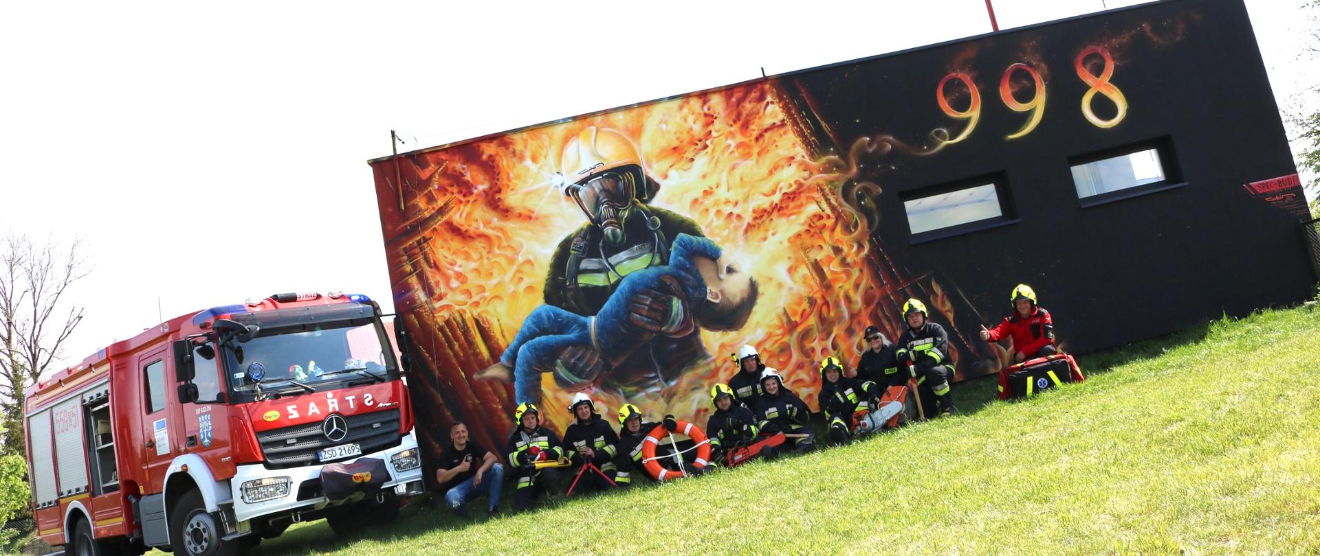 Zdjęcie przedstawia ścianę budynku remizy strażackiej na której jest namalowany strażaka niosący małe dziecko oraz numer alarmowy 998. Przed ścianą siedzą na trawie ochotnicy ubrani w stroje strażacki oraz hełmy strażackiej. Po lewej stronie stoi wóz strażacki marki Mercedes-Benz.