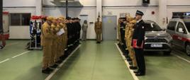 Zdjęcie przedstawia funkcjonariuszy PSP stojących w dwóch rzędach podczas uroczystego zdania służby. Ubrani są w umundurowanie koszarowe koloru pisakowego oraz czarnego. Zdjęcie wykonane w garażu JRG.