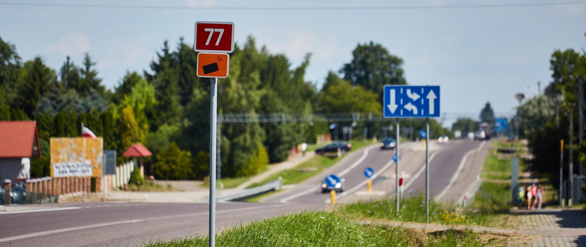 Na pierwszym planie słupek z numerem drogi. Tło rozmyte z zarysem drogi, pasa zieleni, chodnika. Świeci słońce. Widać także niewyraźne samochody na drodze. 