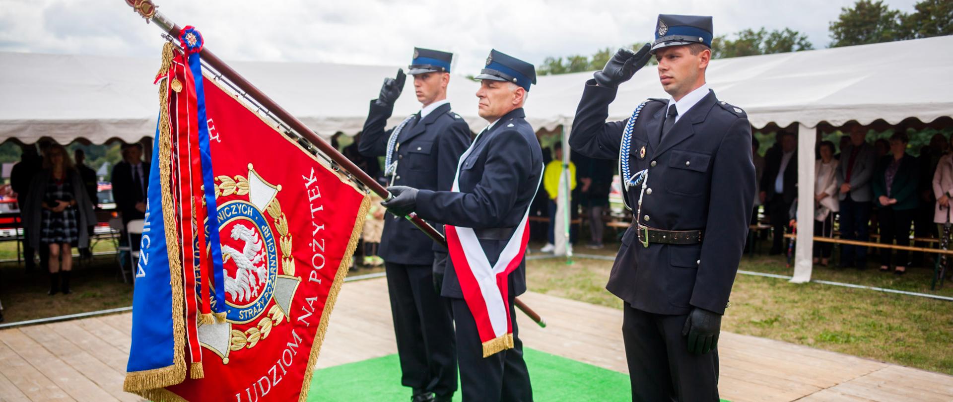 Zdjęcie przedstawia poczet sztandarowy Ochotniczej Straży Pożarnej w Markowej. Trzech strażaków ubranych w ubiory wyjściowe stoją na zielonym dywanie sztucznej trawy. Strażak stojący w środku trzyma sztandar pochylony do przodu. Strażacy po bokach stoją na baczność i salutują