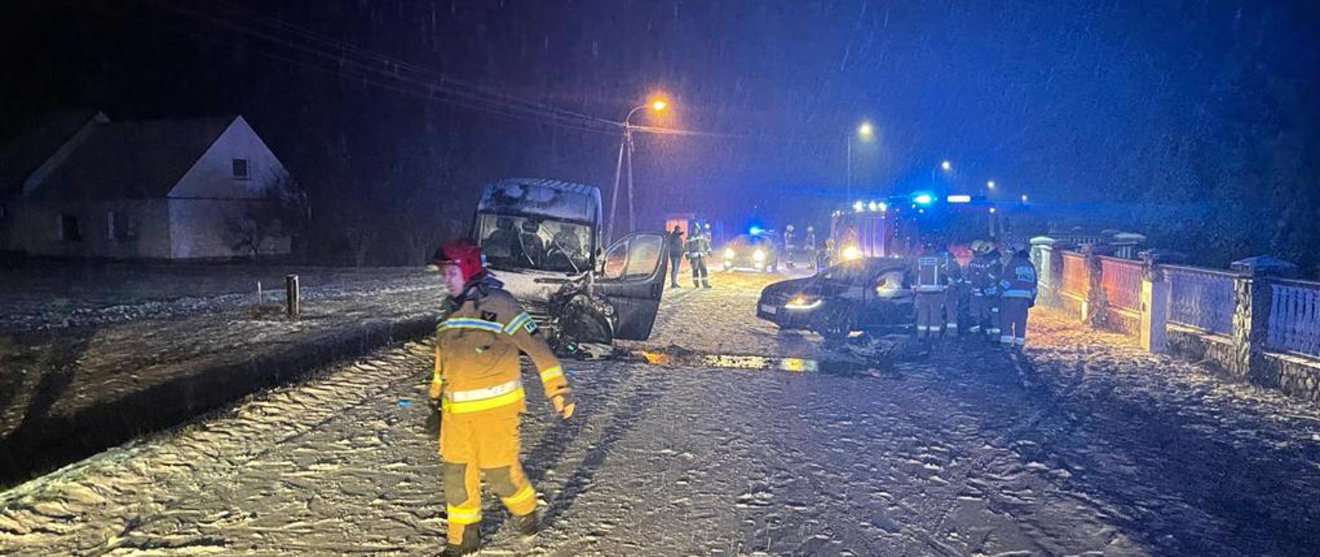 Na zdjęciu widać dwa rozbite samochody, które brały udział w zdarzeniu. Jest pora nocna w tle widać zapalone latarnie drogowe droga jest ośnieżona. Za uszkodzonymi pojazdami widać samochody strażackie