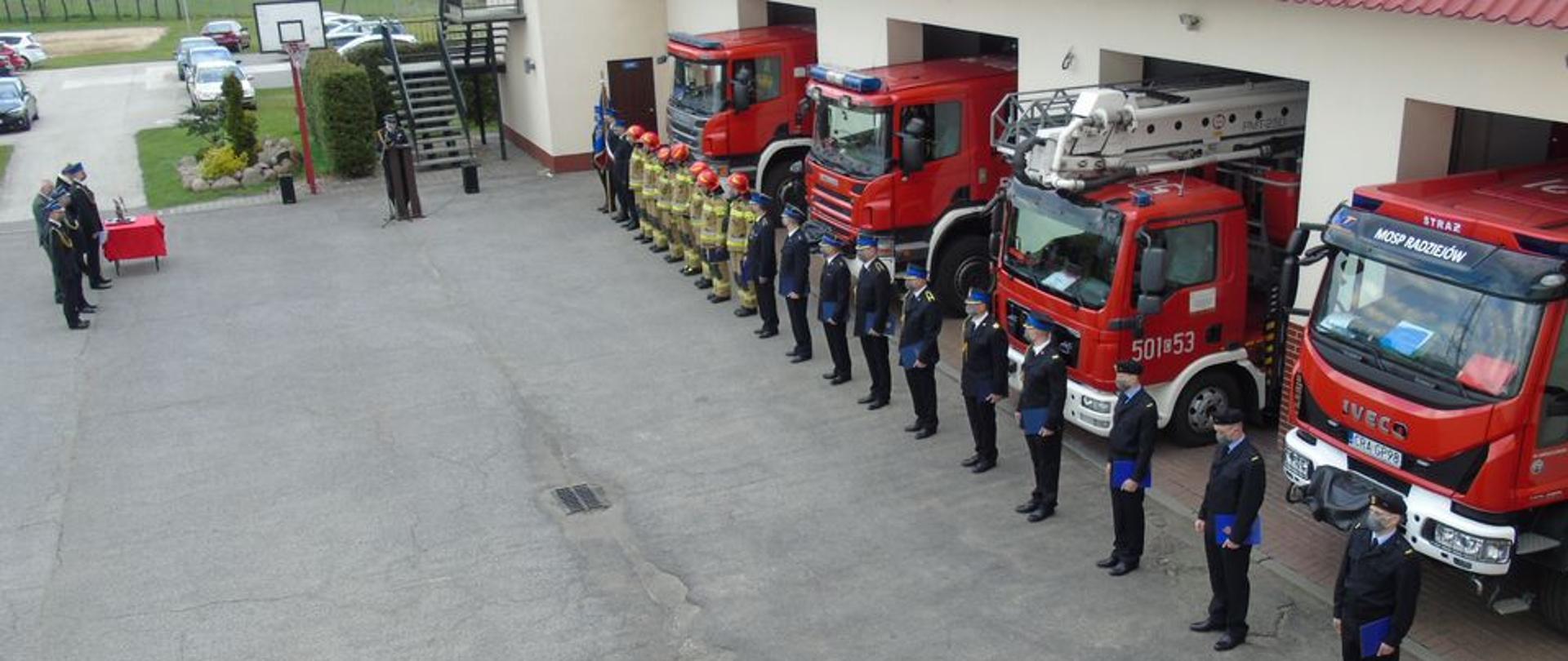 Zdjęcie zrobione z wysokości pierwszego piętra. Na wprost strażak ubrany w umundurowanie wyjściowe ( gabardyna) czapka rogatywka na głowie stoi za drewnianą mównicą, obok mównicy mikrofon na metalowym stojaku. Z tyła zielone krzewy. Po lewej stronie stoi trzech strażaków i dwóch cywili. Po prawej stronie stoją strażacy w szeregu: trzech ubranych w mundury wyjściowe ( gabardyna ), jeden trzyma sztandar, w dalszej kolejności 7 strażaków w piaskowych mundurach bojowych i czerwonych hełmach oraz 10 strażaków w mundurach wyjściowych ( gabardynach) i czapkach rogatywkach. Wszyscy mają założone maseczki. W tle cztery czerwono-szare samochody pożarnicze.