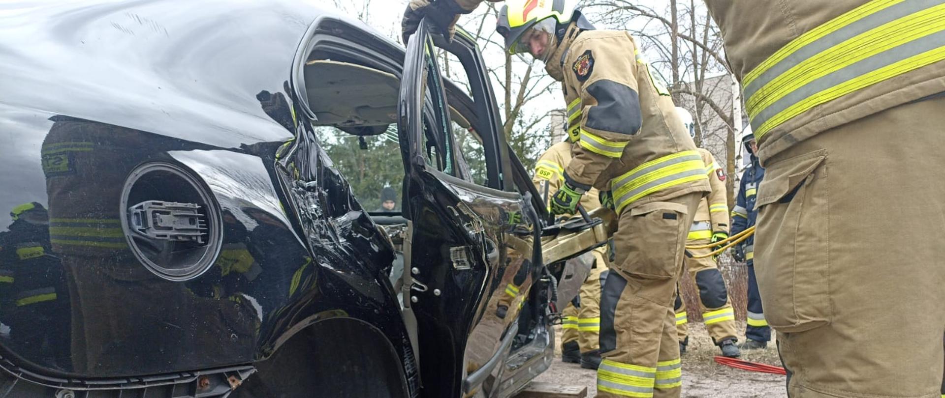 Po lewej stronie zdjęcia znajduje się wrak czarnego samochodu osobowego. Na głównym planie strażak w piaskowym ubraniu specjalnym, trzymając w ręce narzędzie hydrauliczne do ratownictwa stara się wywarzyć drzwi w samochodzie.