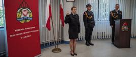Zdjęcie zrobione w pomieszczeniu strażnicy na tle godła Rzeczpospolitej Polskiej, Sztandaru Komendy Powiatowej PSP w Lublińcu, dwóch Flag Rzeczpospolitej Polskiej oraz po lewej stronie banneru Komendy Powiatowej PSP w Lublińcu. Na pierwszym planie nowoprzyjęty strażak stojący na baczność i ubrany w mundur służbowy składa ślubowanie podnosząc prawą rękę, po prawej stronie Komendant w ubraniu galowym stojący za mównicą.