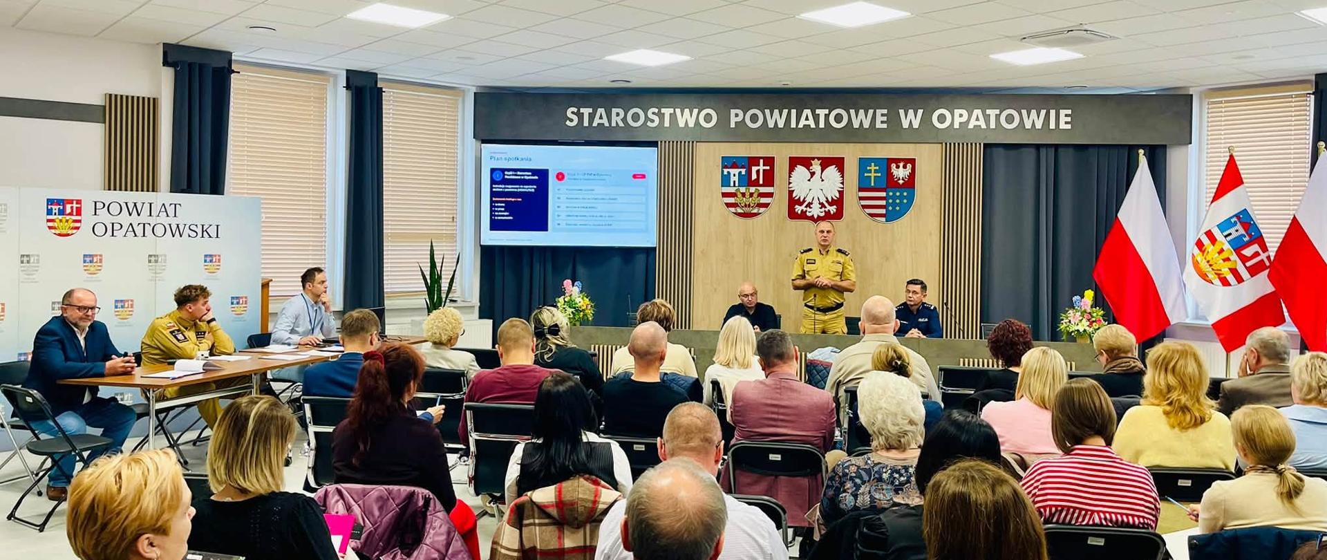 Sala konferencyjna Starostwa Powiatowego w Opatowie. Na pierwszym planie uczestnicy spotkania siedzący na krzesłach, zwróceni w stronę prelegentów. Przy stole oraz na środku sali znajdują się przedstawiciele służb i samorządu. W tle ekran prezentacji, godła i flagi.