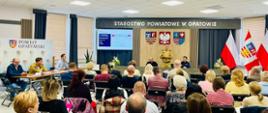 Sala konferencyjna Starostwa Powiatowego w Opatowie. Na pierwszym planie uczestnicy spotkania siedzący na krzesłach, zwróceni w stronę prelegentów. Przy stole oraz na środku sali znajdują się przedstawiciele służb i samorządu. W tle ekran prezentacji, godła i flagi.