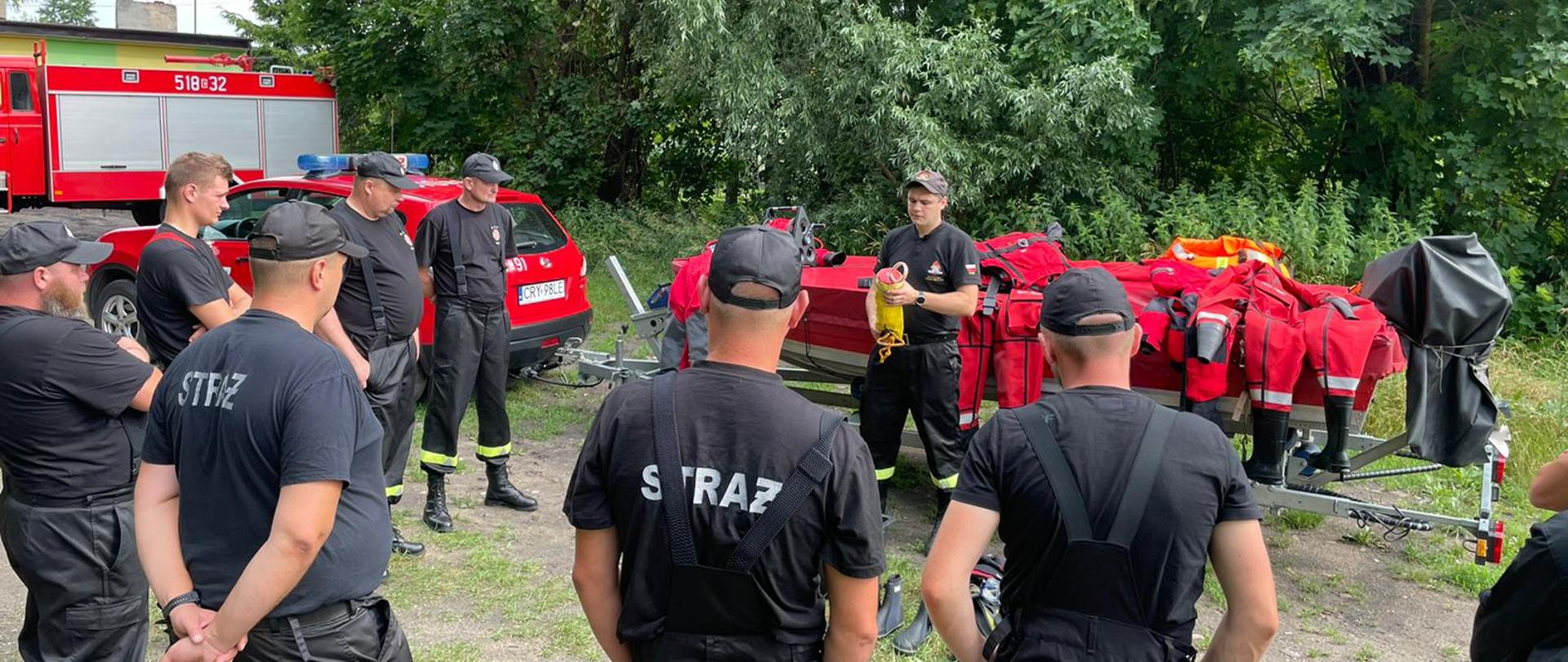 Zdjęcie przedstawia strażaków PSP i OSP podczas ćwiczeń z ratownictwa wodnego. Strażacy ubrani są w ubrania koszarowe. Jeden ze strażaków PSP przedstawia sprzęt wykorzystywany do działań na wodzie. Na zdjęciu widoczne są samochody strażackie oraz łódka. W tle zdjęcia widać drzewa. 