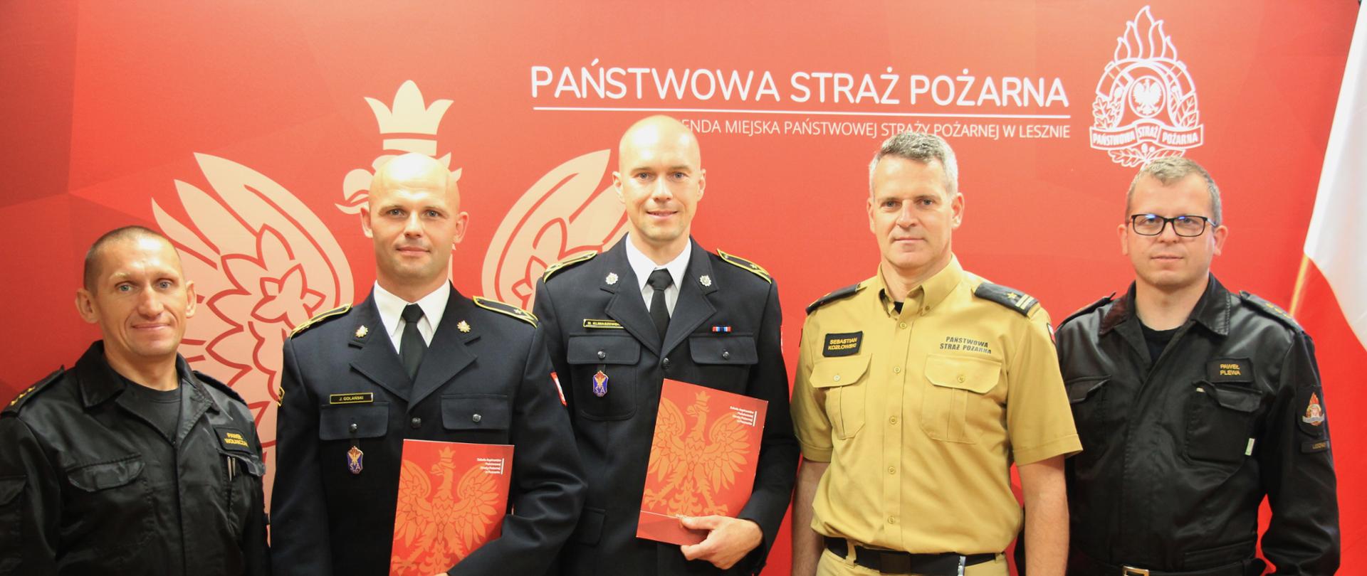 Strażacy w mundurach stoją w szeregu na tle loga PSP.