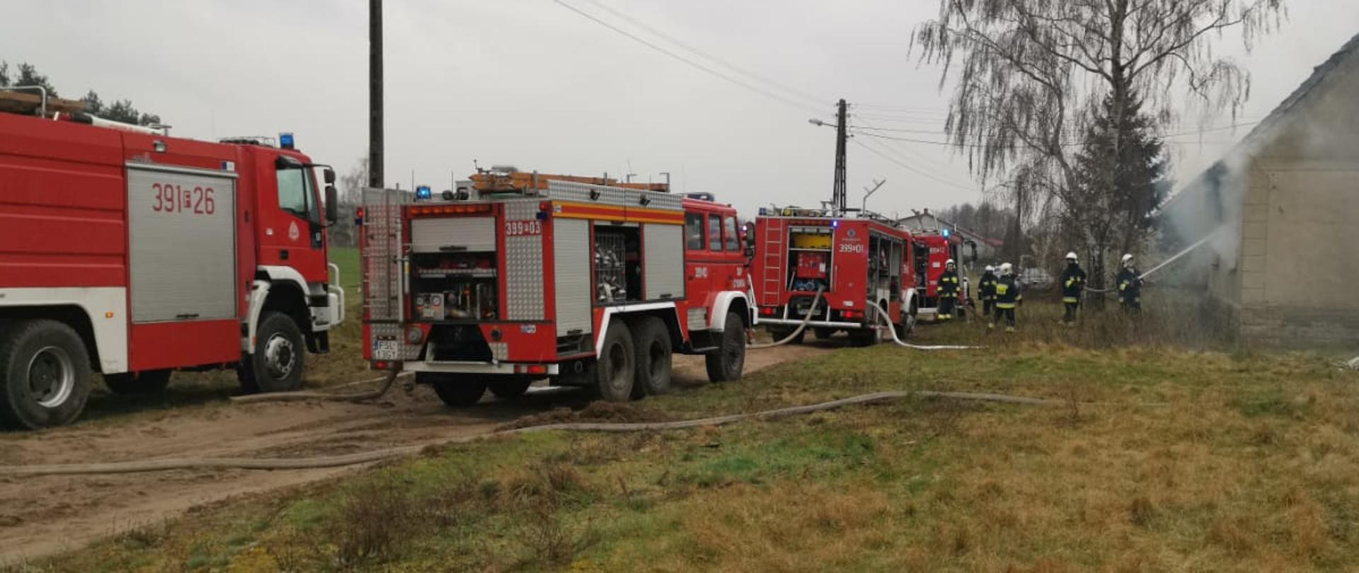 Widok stojących samochodów gaśniczych z rozwiniętymi liniami wężowymi. Po prawej stronie przed zadymionym budynkiem strażacy, podający prąd gaśniczy z zewnątrz przez okno budynku.