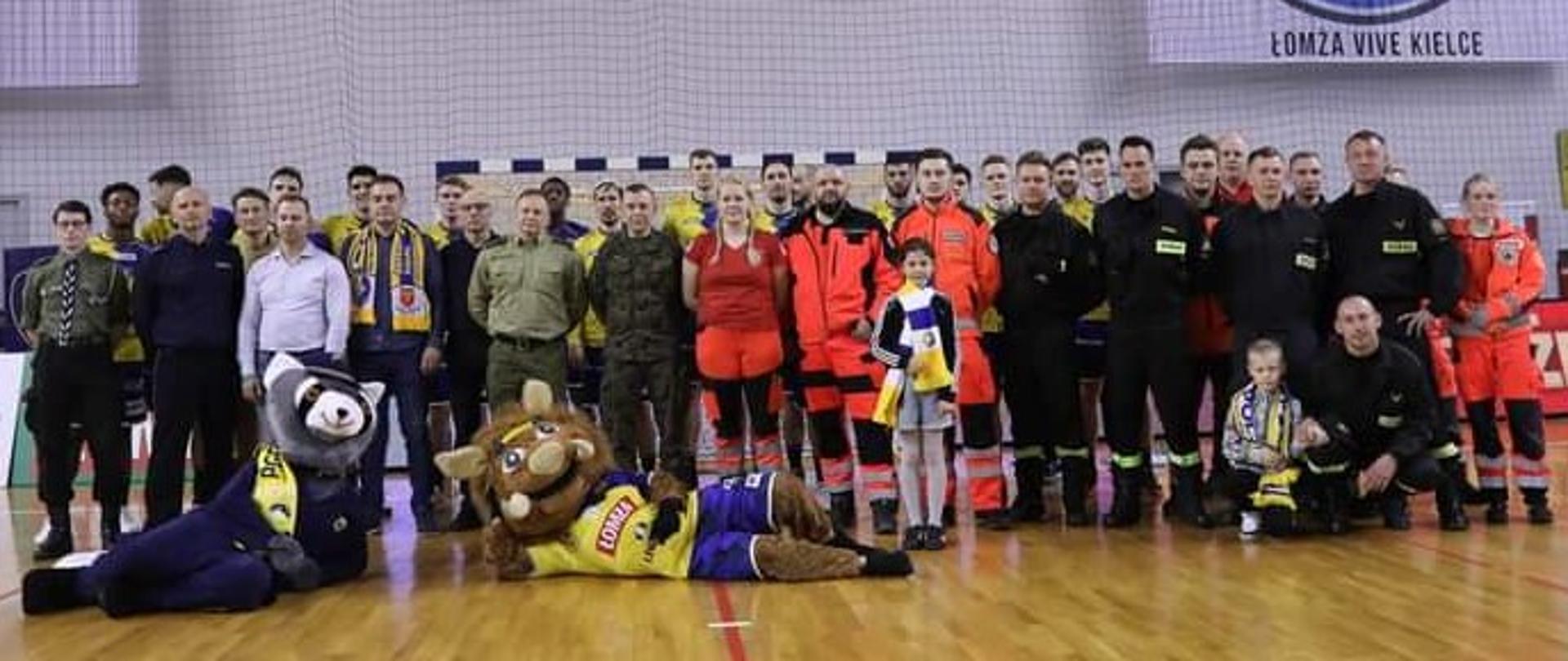 Zdjęcie przedstawia zgromadzonych na meczu przedstawicieli zaproszonych służb mundurowych, medycznych oraz wolontariuszy wraz z maskotkami zespołów sportowych.