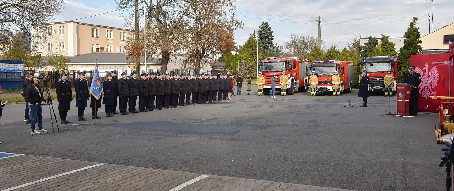 plac przed komendą straży w kutnie na placu po lewej stoi pododdział strażaków PSP po prawej w głębi widać trzy samochody ratowniczo-gaśnicze, w tle budynki i drzewa