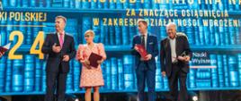 Na tle wielkiego ekranu z napisem Nagrody ministra stoi minister Wieczorek, wiceminister Mrówczyńska i dwóch mężczyzn w garniturach, trzymających czerwone teczki.