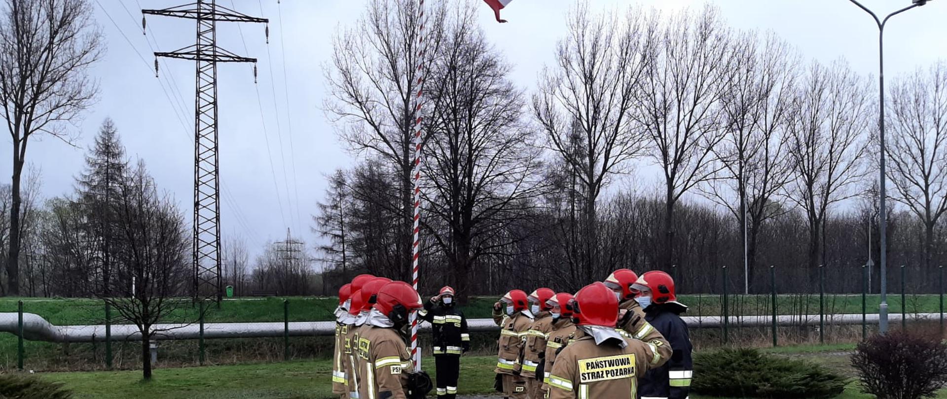 Na placu Komendy Powiatowej Państwowej Straży Pożarnej w Kamiennej Górze odbywa się uroczysta zmiana służby z okazji Dnia Flagi Rzeczpospolitej. Funkcjonariusze w ubraniach specjalnych oddają honory Fladze państwowej. Dowódcy stoją na przeciw siebie z tyłu widoczny masz w kolorze biało czerwonym na którym powiewa flaga państwowa, 