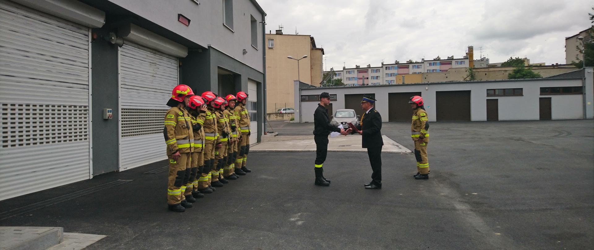 Po lewej stronie strażacy ustawieni w szeregu. Przed szeregiem nowy strażak któremu Komendant Powiatowy wręcza akt mianowania.