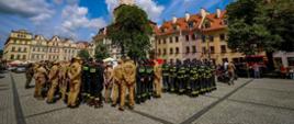 Szerokie ujęcie przedstawiające formacje strażackie zebrane na brukowanym rynku w Jeleniej Górze. Strażacy w piaskowych oraz ciemnych mundurach stoją w dwu rzędach, zwróceni tyłem do obiektywu. Nad placem góruje wysoka wieża ratuszowa z zielonym dachem, otoczona zabytkowymi kamienicami z pomarańczowymi dachami. Nad rynkiem rozciąga się błękitne niebo z dynamicznie ułożonymi, białymi chmurami