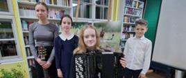 Zdjęcie czworo uczniów wykonane w bibliotece na tle półek z książkami. W centrum, siedząc na krześle, znajduje się dziewczynka ubrana na czarno, trzymająca akordeon. Za nią stoją dwie dziewczynki – jedna z nich trzyma gitarę klasyczną, obie ubrane elegancko, w granatowych sukienkach. Po prawej stronie stoi chłopiec w białej koszuli i ciemnych spodniach. Na ścianach widać żółte i zielone elementy wystroju, a z sufitu zwisają linki z oświetleniem. W tle widoczny jest również ekran projekcyjny oraz zdjęcie ptaka. 