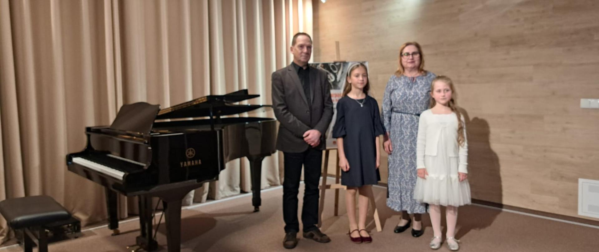Na zdjęciu widoczna jest grupa czterech osób stojących obok czarnego fortepianu Yamaha. Od lewej strony stoi mężczyzna w ciemnym garniturze. Po jego prawej stronie stoi dziewczynka w ciemnej sukience. Następnie stoi kobieta w okularach i w jasnej sukience we wzory. Obok niej stoi dziewczynka w jasnej sukience. W tle widoczne są jasne zasłony i drewniana ściana. Na dole zdjęcia widnieje napis "12 maja 2025".