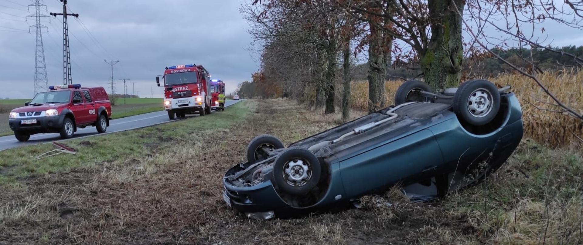 Na zdjęciu samochód osobowy leżący w rowie kołami do góry (na dachu), obok na jezdni stoją 3 pojazdy pożarnicze z włączonymi sygnałami świetlnymi.