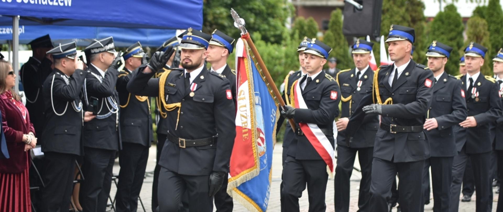 Defilada strażacka na placu miejskim. Na przedzie maszeruje poczet sztandarowy – trzej strażacy, z których środkowy niesie dwustronny sztandar z napisem „W SŁUŻBIE...”. Strażak po lewej stronie salutuje. Kolumna przechodzi obok niebieskiego namiotu z napisem „Miasto Sochaczew”, pod którym stoją zaproszeni goście.
